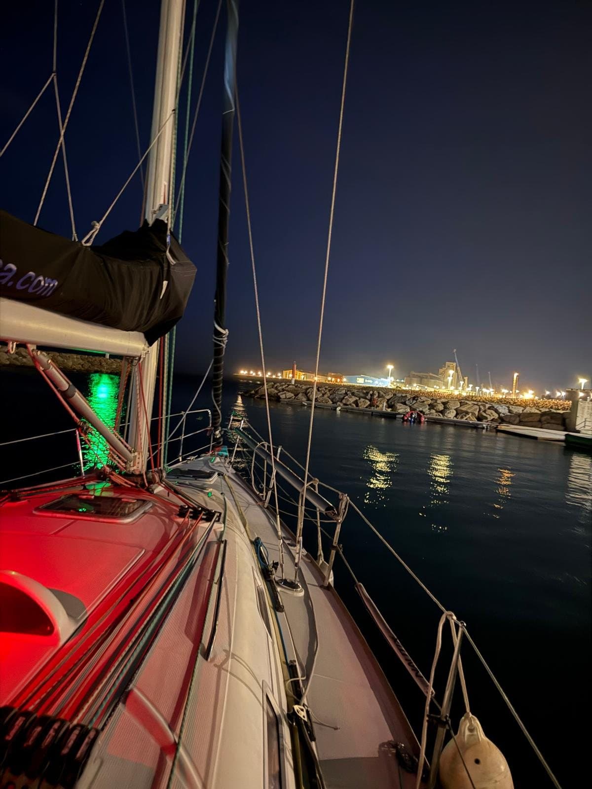 Proa de un velero de noche con las luces de navegación encendidas frente a un puerto iluminado